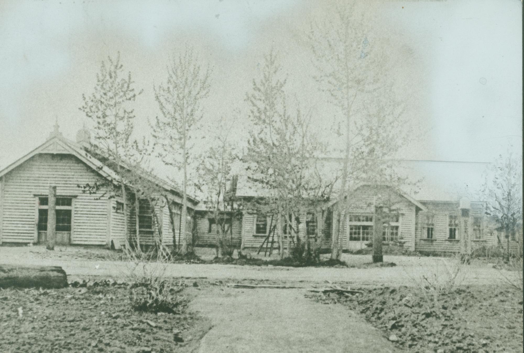 手前に数本の木が横一列に並んで立ち、L字型に造られた1階建ての校舎の白黒写真