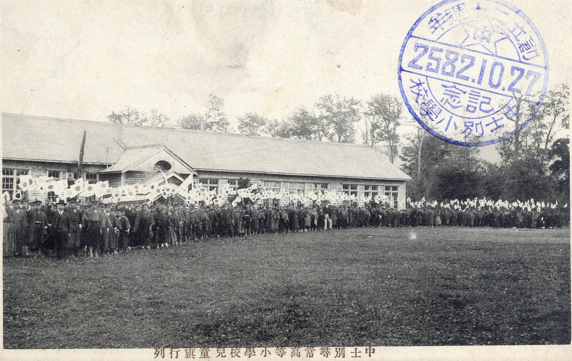 校舎の前に大勢の人達が集まり日の丸の旗を振っている大正時代の中士別小学校校舎の白黒写真