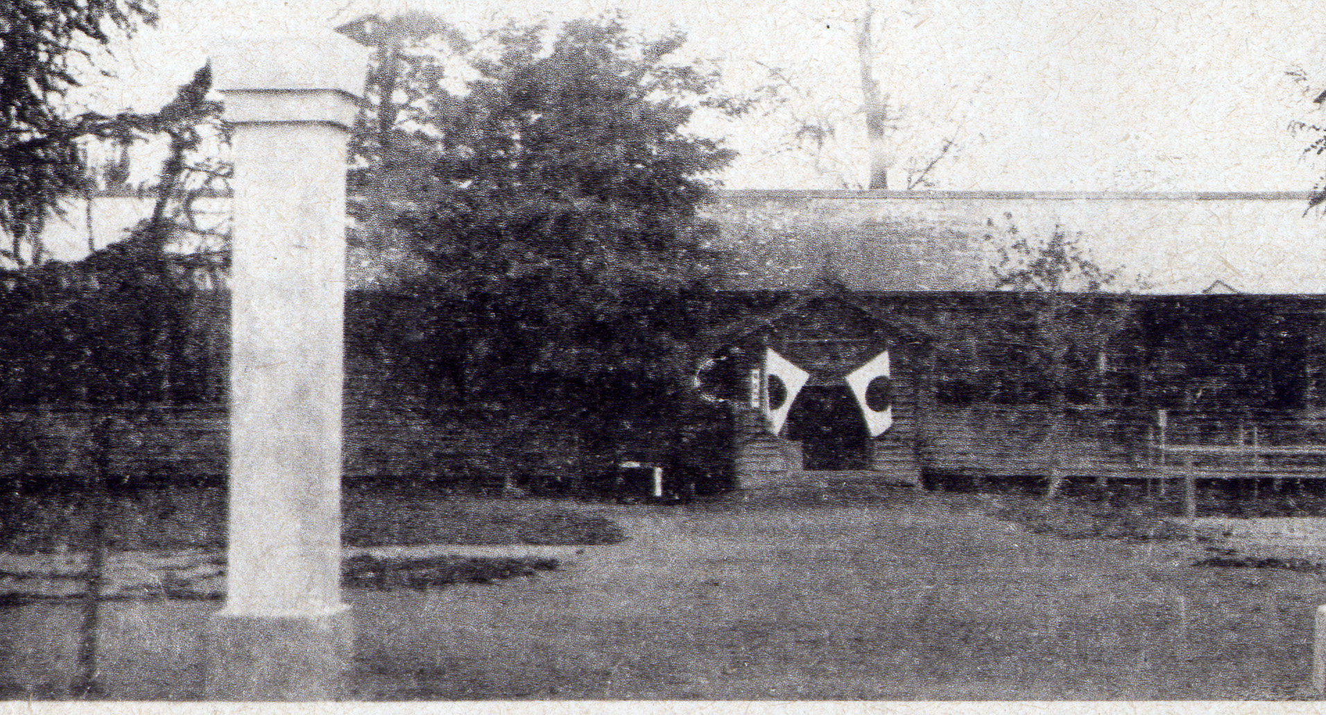 大きな門柱が立つ奥の校舎の入り口に2つの日の丸旗が飾られている1階建ての大正時代の多寄小学校の校舎の写真