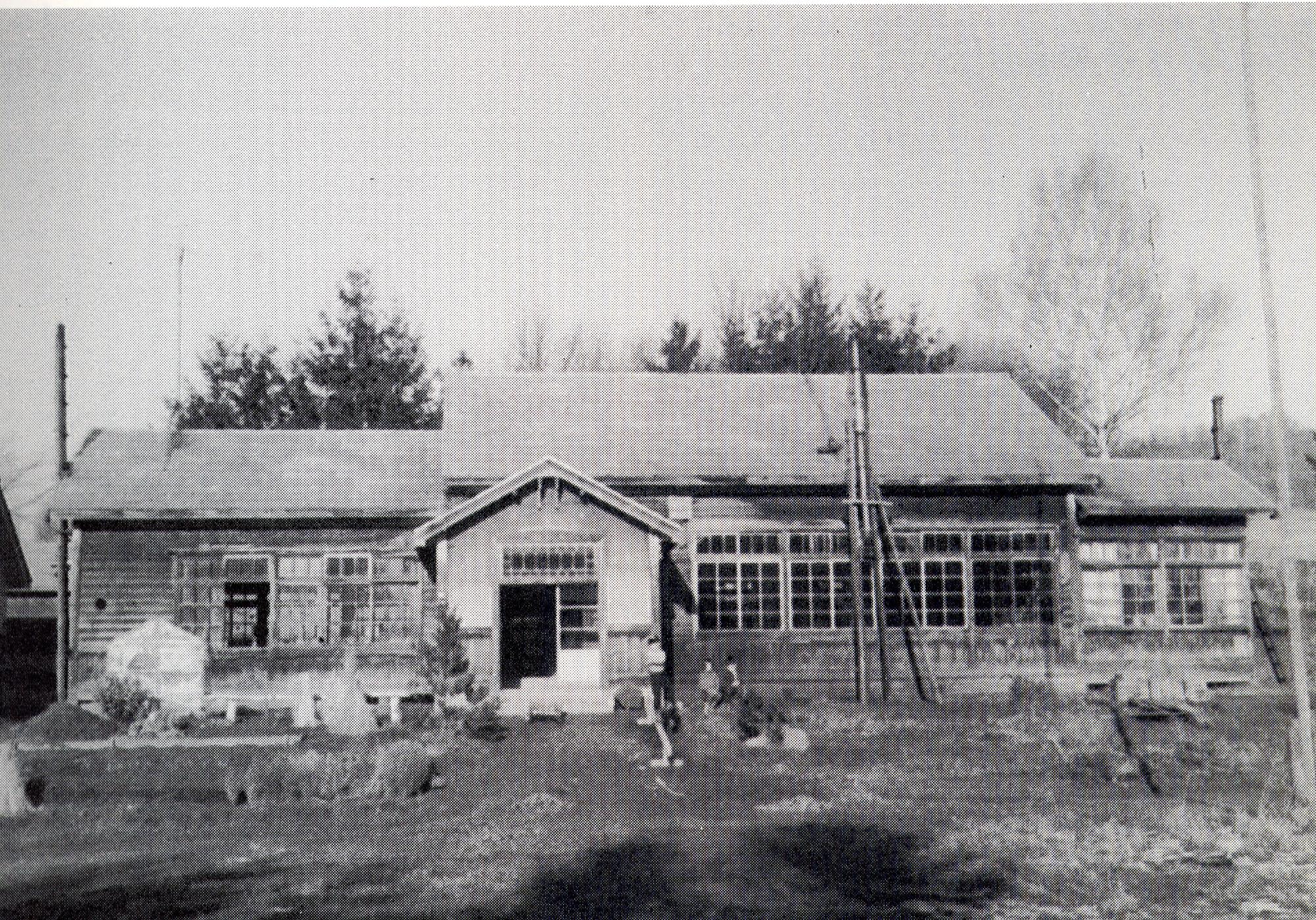 中央入口の右側に1名の児童が立ち、隣に2名の児童が座っている旧三郷小学校校舎の白黒写真
