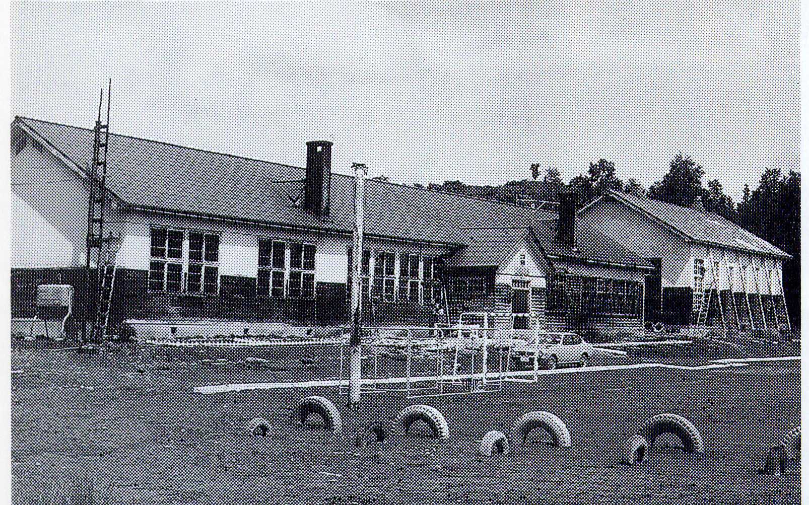 屋根の左右に煙突が設置された旧北温小学校校舎をタイヤ遊具が設置された校庭から撮影した白黒写真