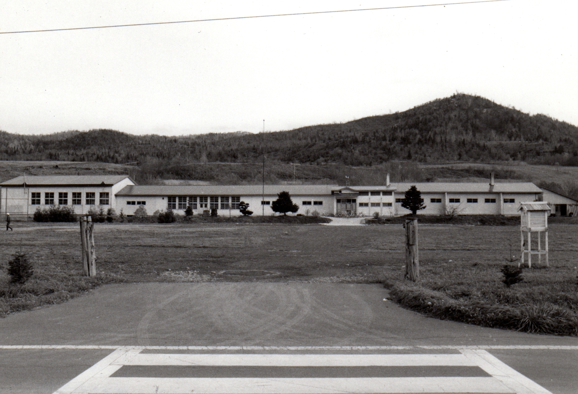 小山の麓に建てられた広い校庭の奥に横長の旧茂志利小学校校舎を道路側から撮影した写真