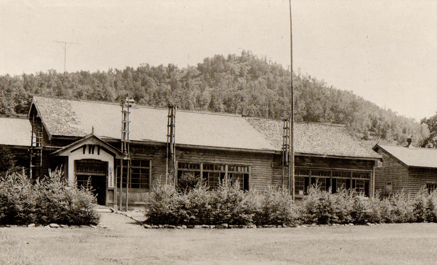小山の麓に建てられ手前に生け垣がある左側に入り口が設けられた昭和37年頃の旧茂志利小学校校舎の白黒写真