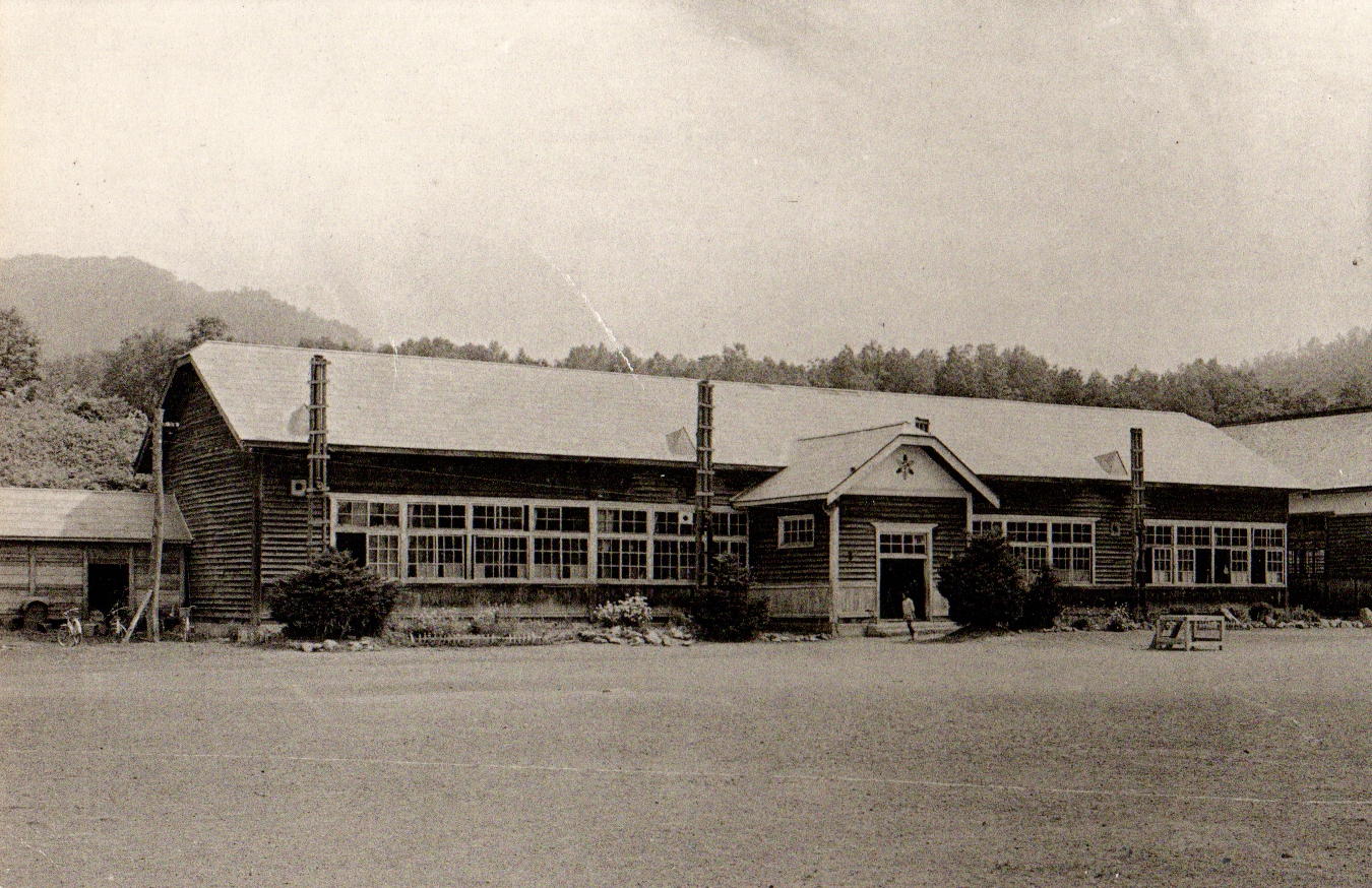 林の手前に建てられた旧似峡小学校校舎の入り口の前に1名の児童が立っている白黒写真