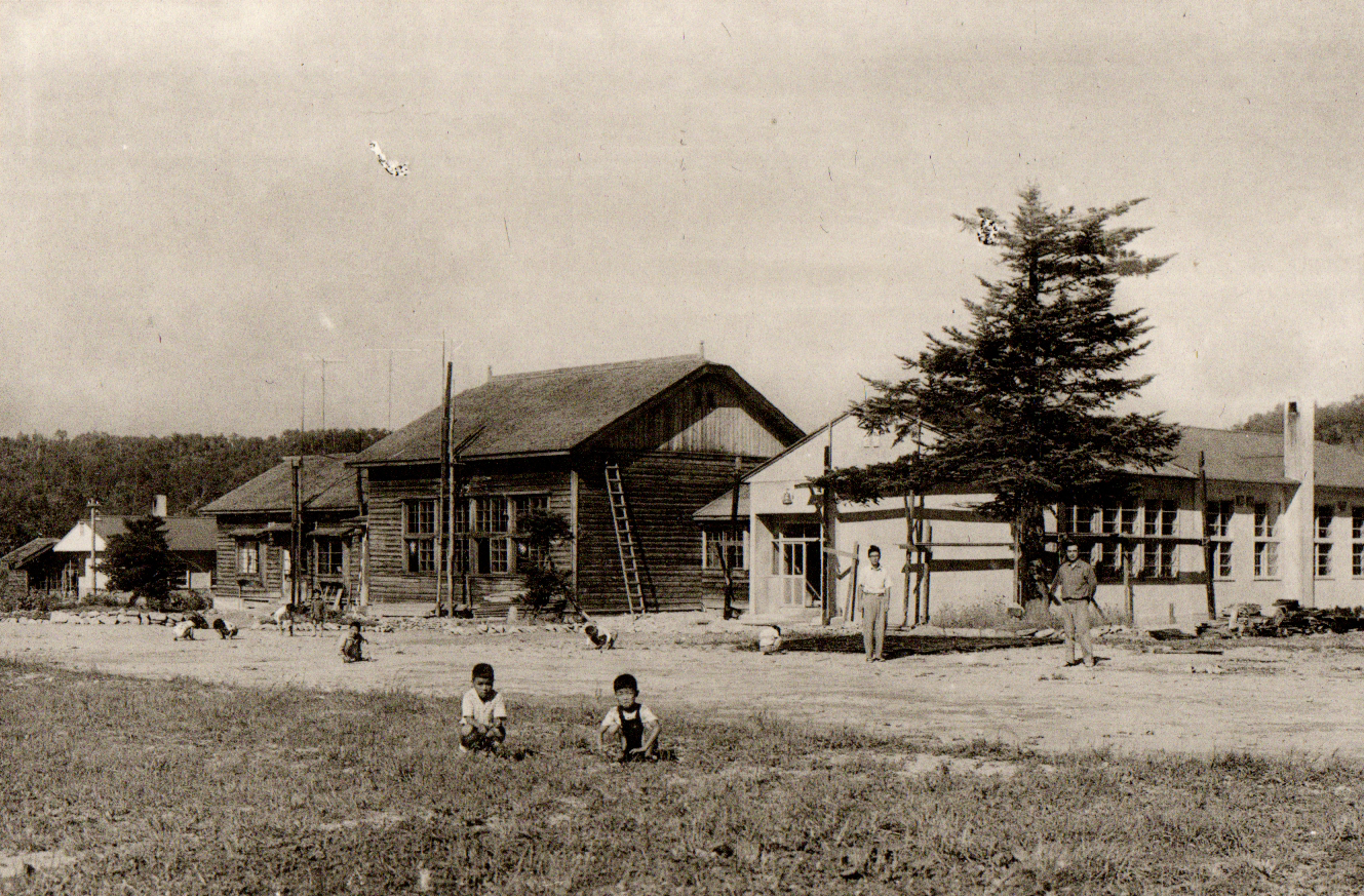 旧上似小学校校舎の前の1本の木の前で2名の男性が間隔をあけて立ち、手前の広場で児童が座って遊んでいる写真