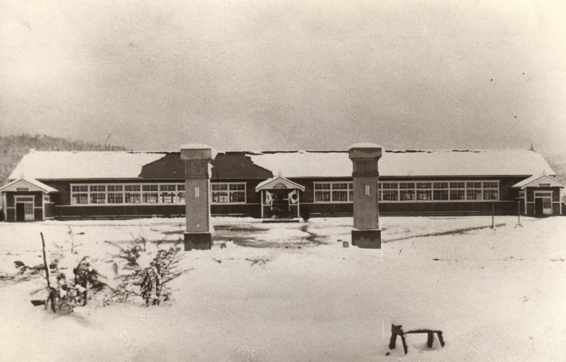 中央に大きな2つの門柱が立ち、奥の入り口に日の丸旗が飾られている雪が積もった昭和3年時の横長の糸魚小学校校舎の白黒写真