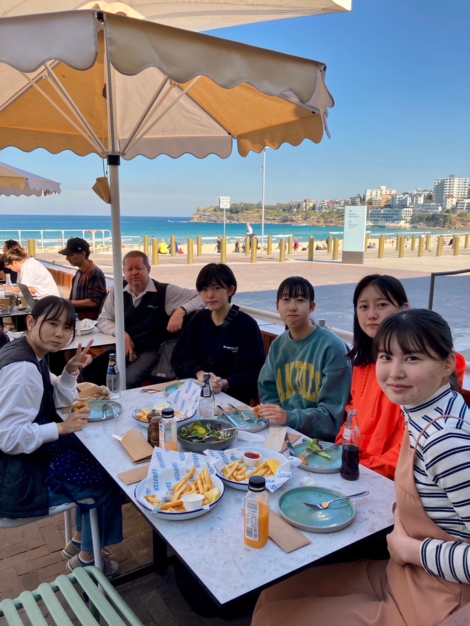 ビーチ沿いのテラス席で食事をする生徒5人とGM市担当者の写真