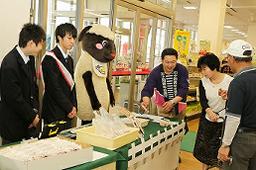 制服を着た2名の男子学生とマスコットキャラクターのみーちゃんが販売ブースに並んで立ち、商品を見ている夫婦に男性が話しかけている写真