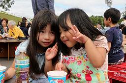 祭り会場でかき氷を食べている2名の女の子が寄り添い、右側の女の子がカメラに向かってピースサインをしている写真