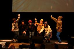 ステージ上に、ニット帽をかぶった6名の女性が集まり両手を広げている写真