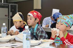 三角巾とエプロンを着用した3名の男の子が美味しそうにおにぎりを食べている写真