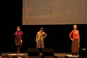 ステージ上に3名の女性が横一列に立ちポーズを取って立っているファッションショーの様子の写真