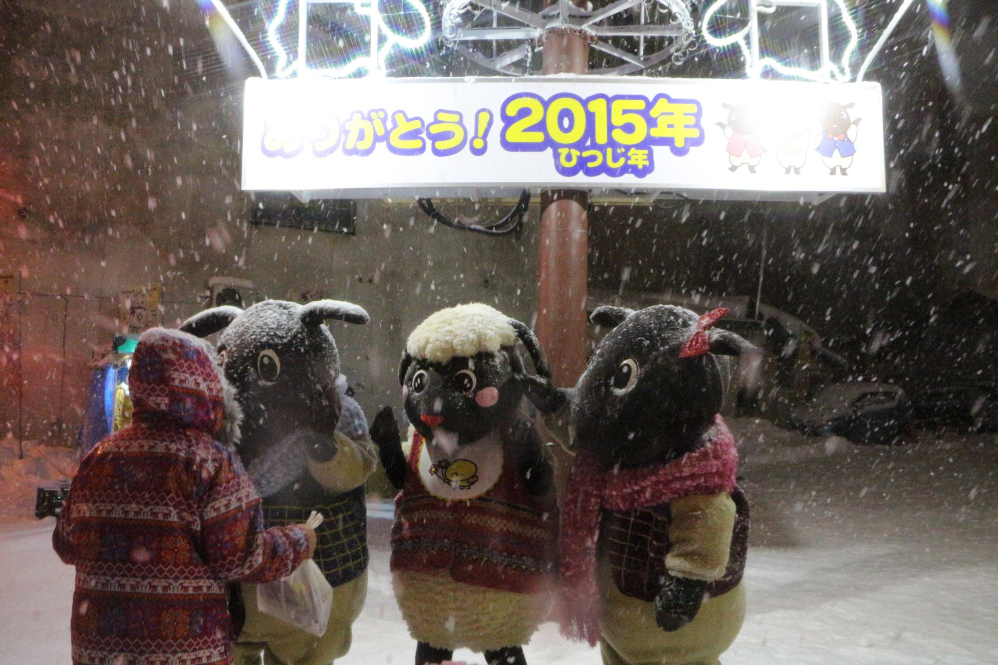 雪が降るなか、「ありがとう！2015年ひつじ年」と書かれた看板の下に立っている3体のマスコットキャラクターとフードを被った人が向かい合っている写真