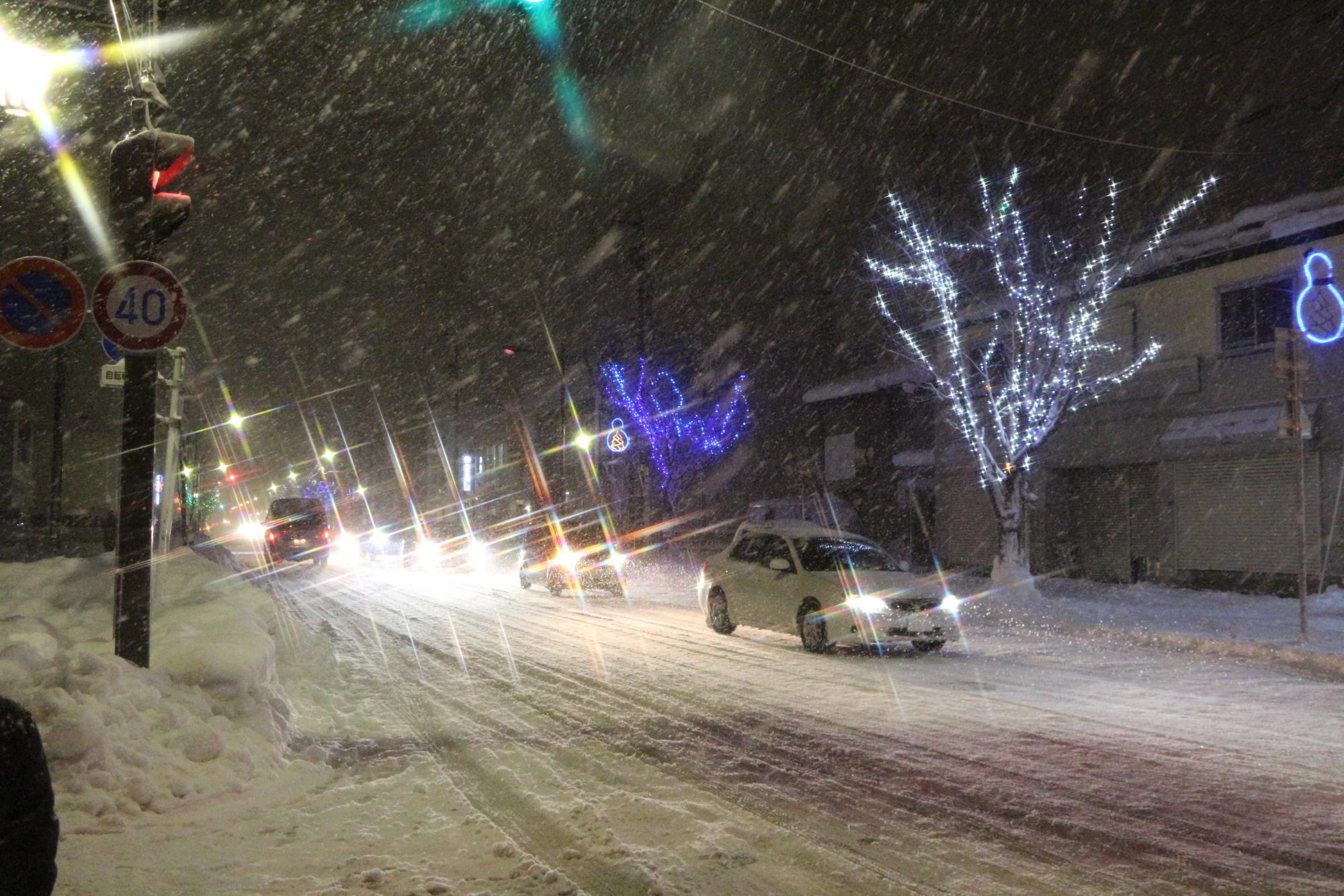 雪が降り積もるなか、街路樹に装飾された紫色や白色に光るイルミネーションの通りを車が走行している様子の写真