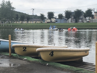 手前に3艘の手漕きボートが置かれた池の奥でペダルボートやアクアサイクルに乗り楽しんでいる人たちの写真