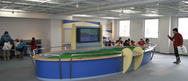 展示室で大型ジオラマを見学しながら職員の話を聞いている子供達の写真