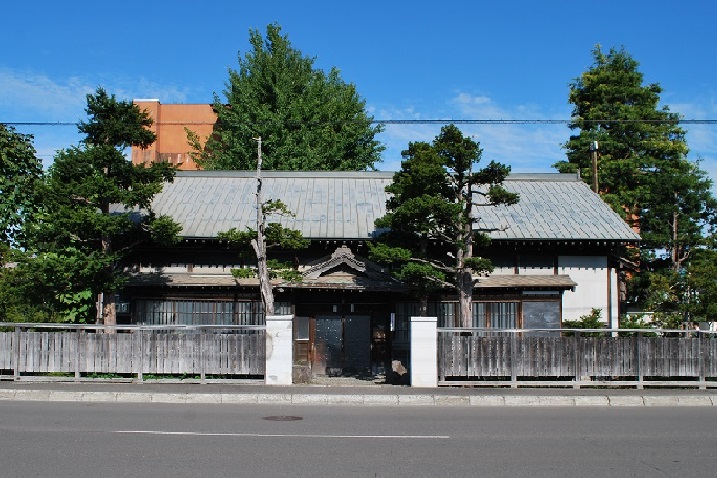 道路に面した場所で垣根で囲われた平屋づくりの日本家屋の建物と周囲に立っている背の高い木々の写真