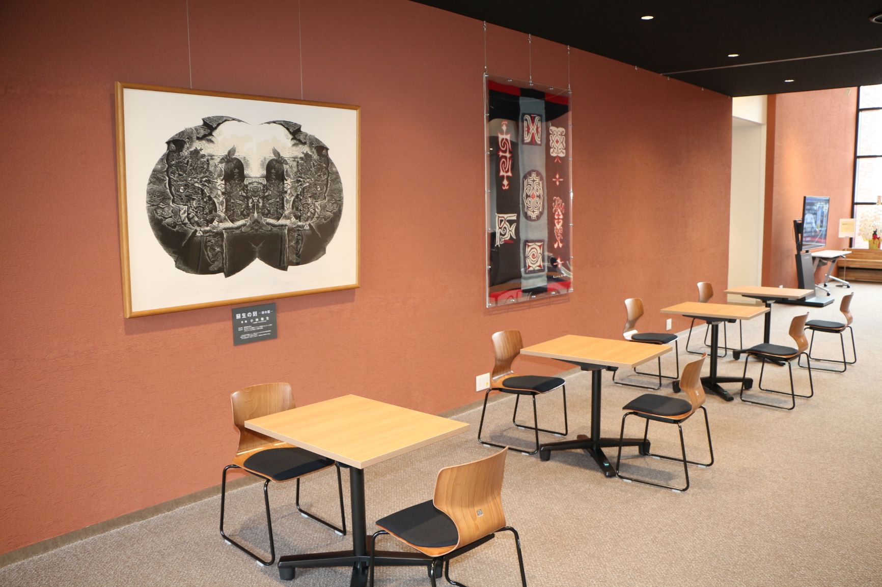 壁に大きな絵画などが展示され、手前に2人掛けの椅子が設置されている木製テーブルが4つ設置された市民テラスの写真