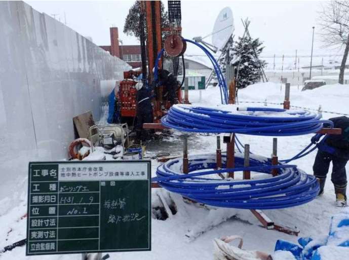 雪が降る中、丸く巻かれた青色のホースが2段に積まれ、奥にある大きな機械につながっている地中熱ヒートポンプ設備導入工事の様子の写真