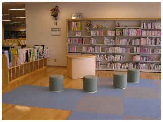 壁に設置された棚に本が収納されており、カーキー色の丸い椅子が4つ、1台の机が置かれた読み聞かせ室の写真
