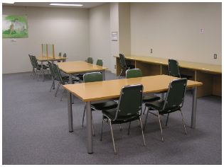 4脚ずつ椅子がセットされた3台の木製テーブルが並び、壁側に長机が設置された学習室の写真