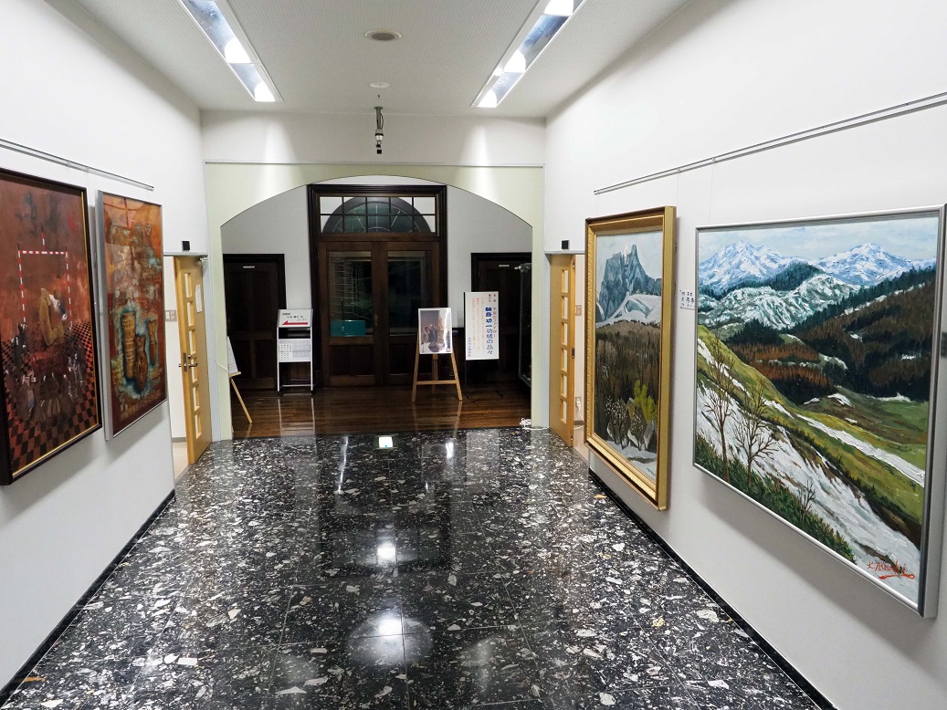 大理石調の床に両サイドの壁に大きな絵が飾られ、奥に入口が見える公会堂展示館の内部の写真