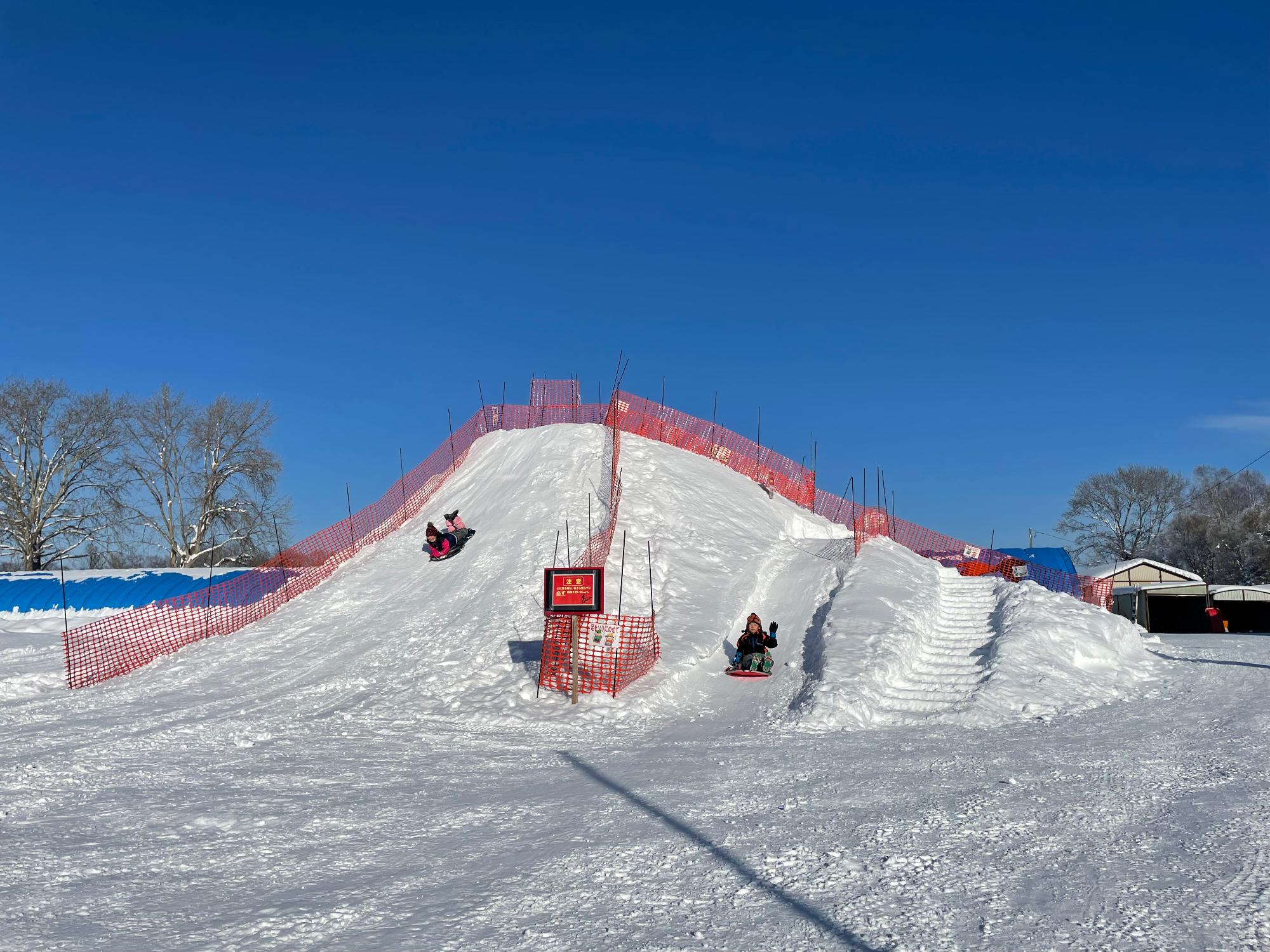 青空の下、雪を高く積み上げた滑り台でそり滑りを楽しんでいる人たちの写真