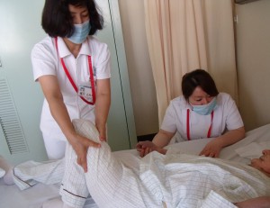 一人の看護師がベットに寝ている患者さんの左脚を立てて、もう一人の看護師が右手を握り、動きの支援を行っている様子の写真
