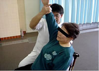 座っている女性が右手を上げ、療法士の男性が右手をつかんで支えている写真