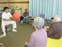 前に座る男性が大きなボールを持ち、対面に座っている高齢者の女性2人がそれを見ている様子の写真