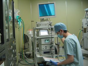 手術着を身に着けた男性が、機械の前に座り、機械と手元の資料を見ながら作業をしている様子の写真