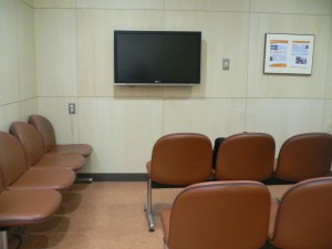 正面壁にテレビが掛けられ、左側に茶色い4人掛けの椅子と、中央に3人掛けの椅子が2つ置かれている待合室の写真