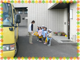 停車している黄色い通園バスの横で、保育士の前に帽子とカバンを持った園児5名が並んでいる写真