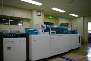 横長の白い機械の上に小さなディスプレイと青色の透明のカバーが設置されている生化学・免疫検査の機器の写真