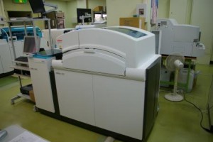 中央の上部が弯曲した透明になっている1メートルくらいの白色の機械、左側にパソコンとキーボードが設置された生化学・免疫検査の機器の写真
