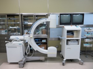左側にCの形をした撮影装置、右側に棚の上に2つのディスプレイが設置されたポータブルX線透視装置の機器の写真