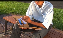 白いワイシャツを着た男性がベンチに座り、冊子本を開いて見ている写真
