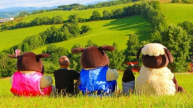 緑が広がる草原に、士別市の3体のマスコットキャラクターが座り、その間に男性と子供が座っている様子を後方から写した写真