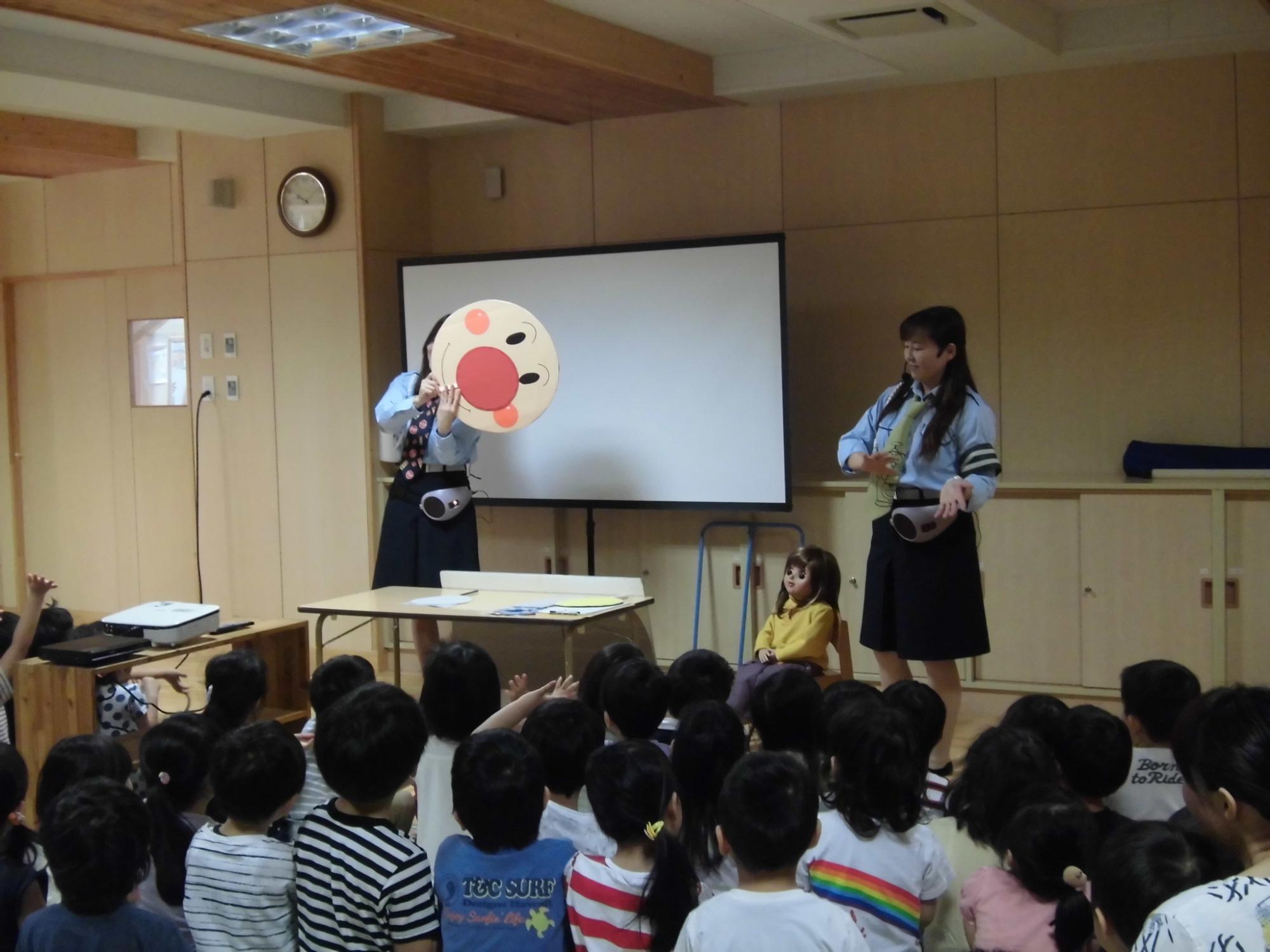 園児たちの前に立ち、アンパンマンの顔やお人形を使って説明している2名の交通安全教育隊を教室の後方から写した写真