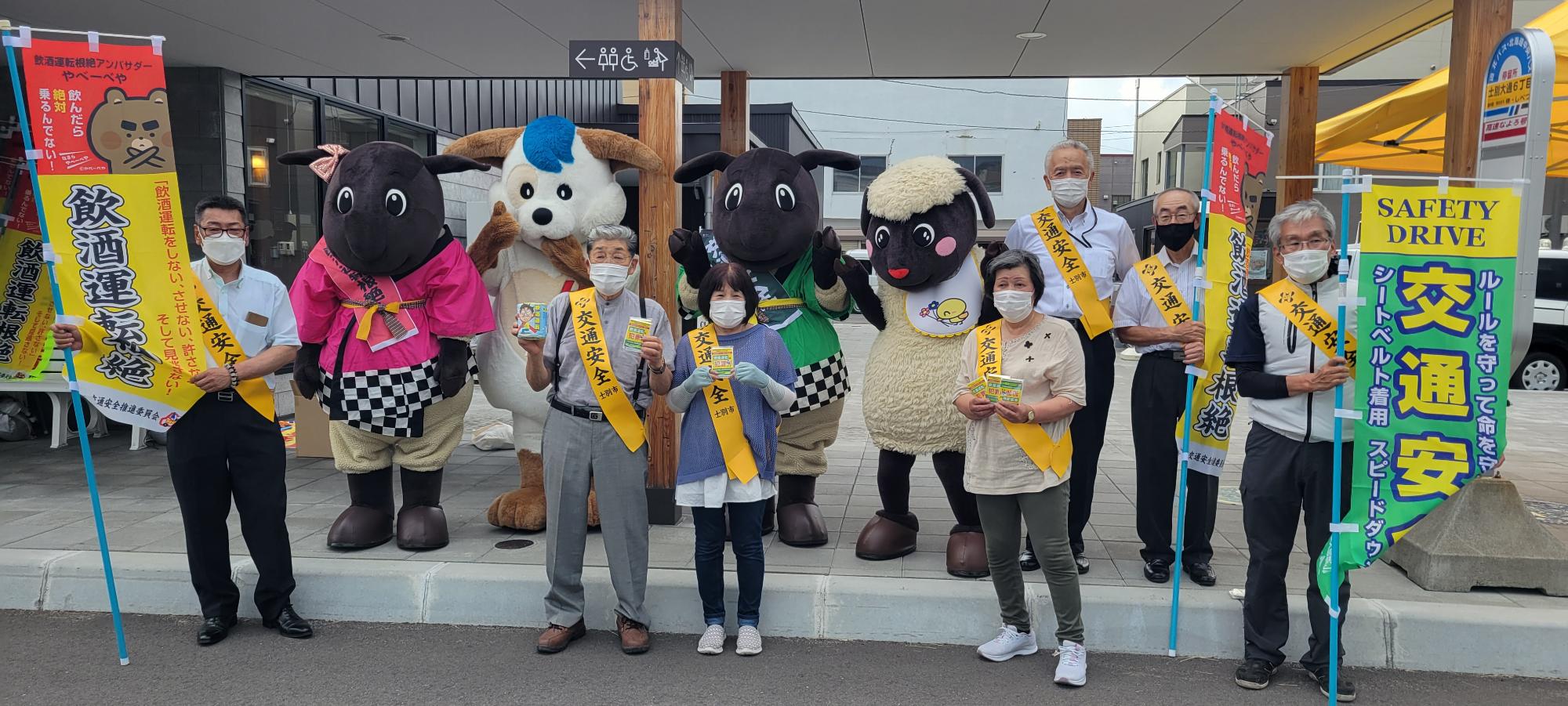バス停留所でさほっちファミリーと北海道警察マスコットのほくとくん、黄色のたすきを身に着けた4名の安全協会の方、のぼり旗を持った3名の男性達が2列に並んでいる集合写真
