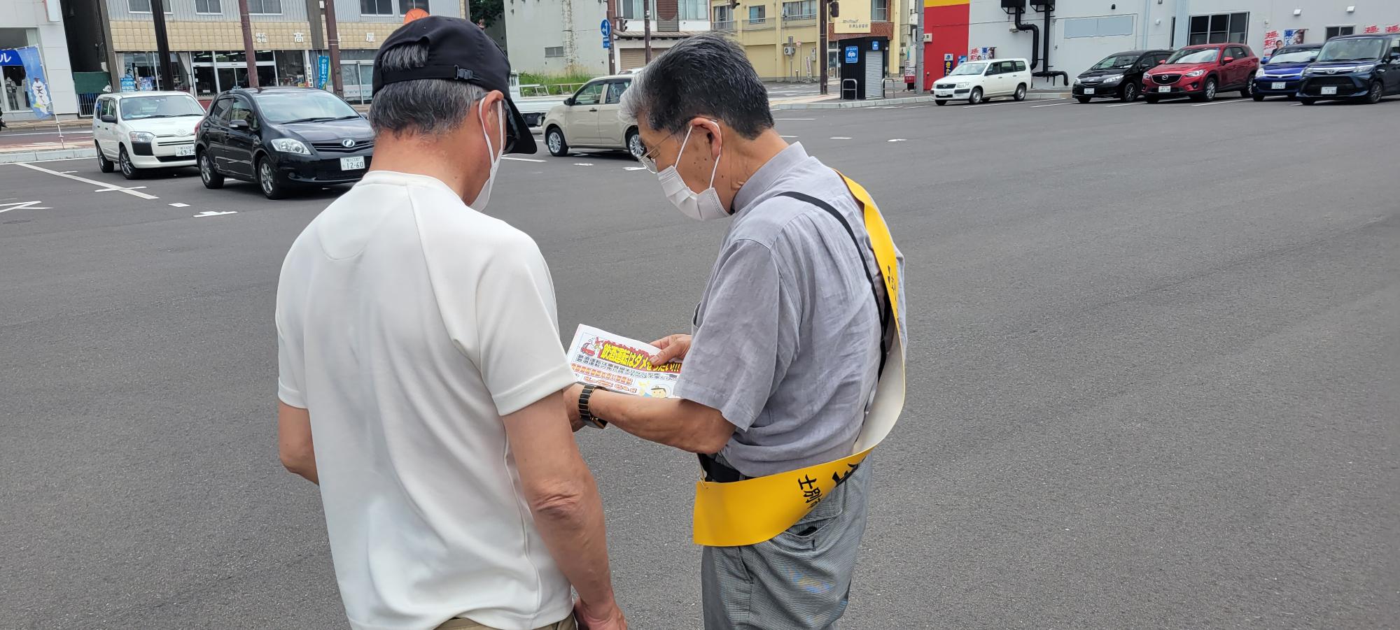 黒色のキャップを被った男性に黄色のたすきを身に着けた安全協会の男性が声をかけて話をしている後ろ姿の写真