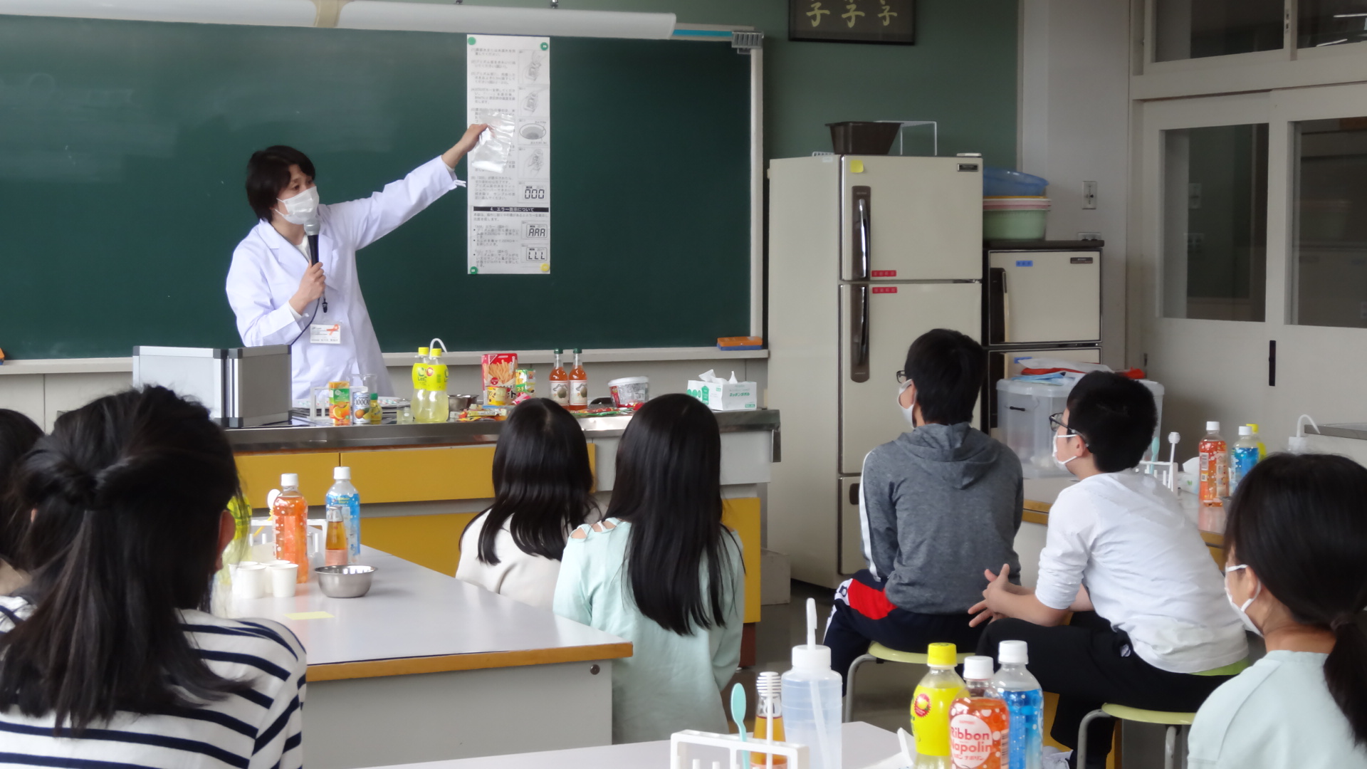白衣を着た女性が黒板に貼ってある資料を見ながら生徒達に説明している授業風景の写真