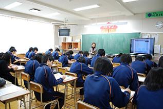 黒板前で話をしている女性と、机に向かいノートなどに書き込んでいる生徒たちの授業風景を教室の後方から写した写真