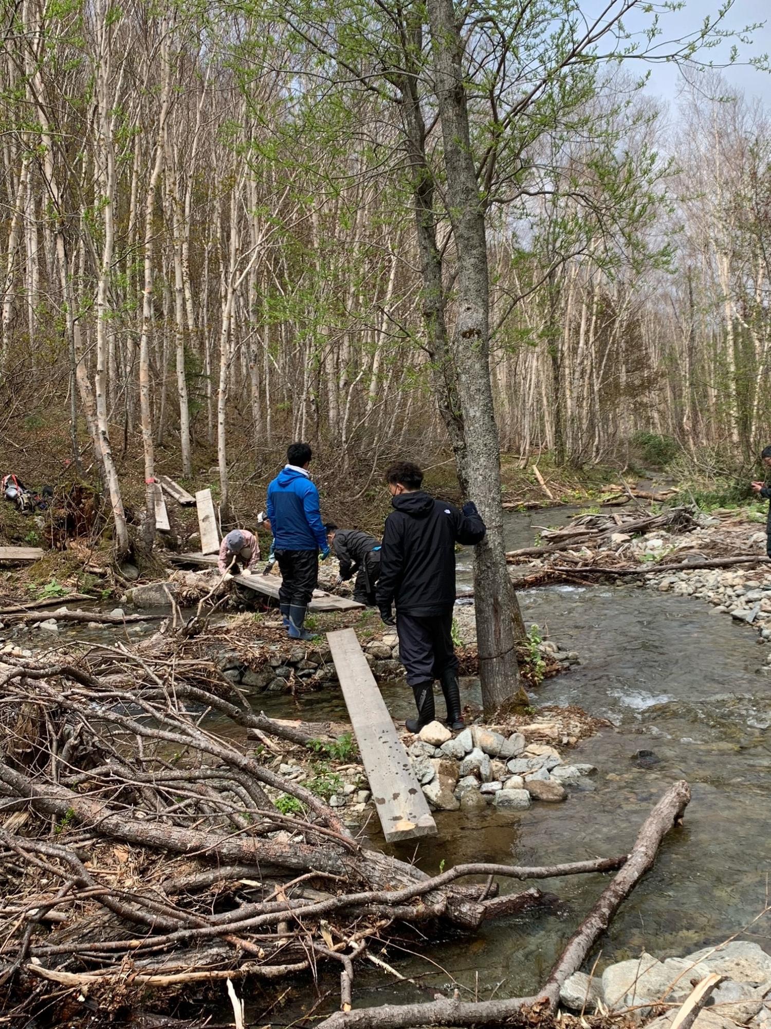 山中の小川で板を組み合わせて橋を作る作業をしている紙谷隊員と朝日支所職員の方々の写真