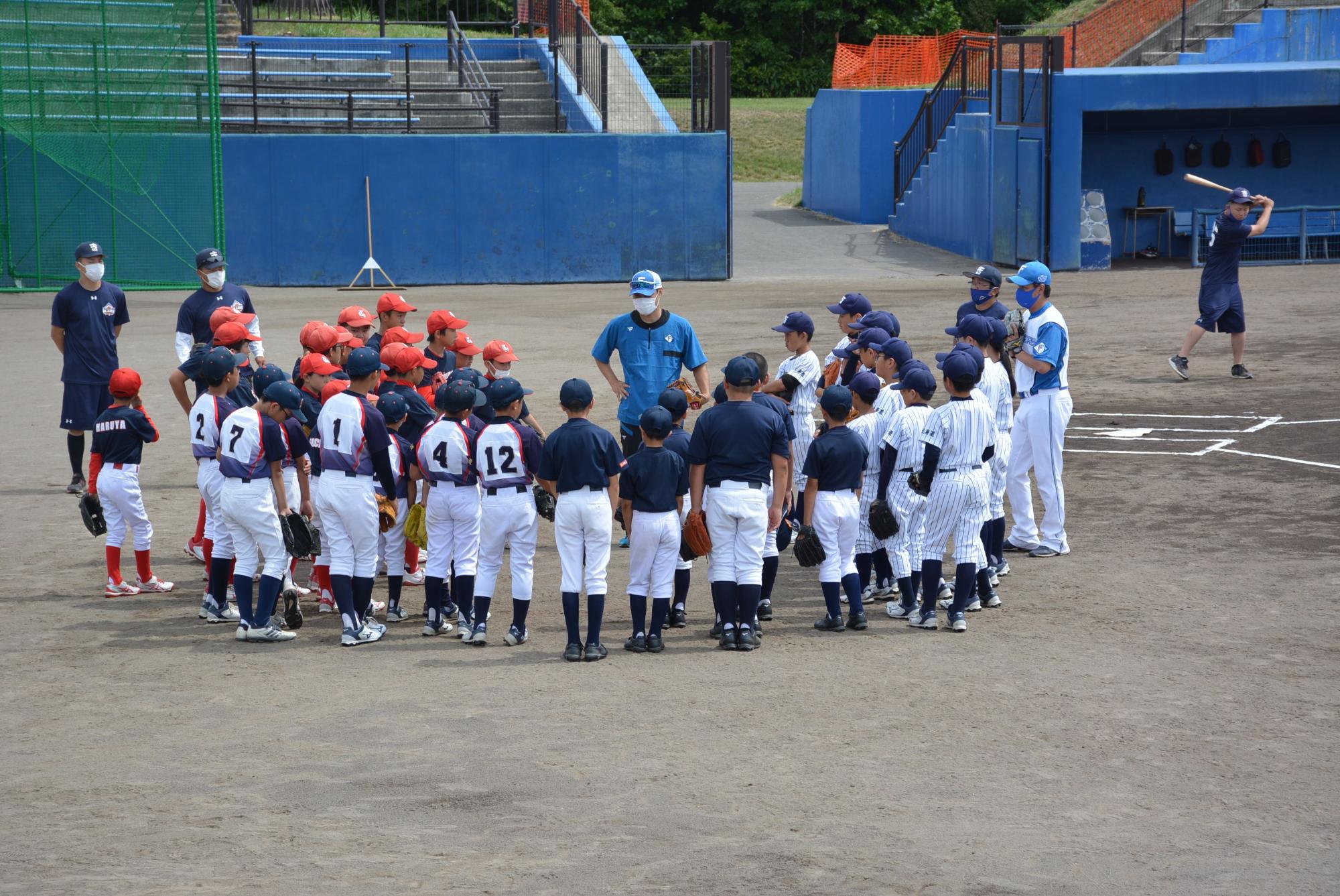 ユニフォームを着た野球少年たちが青色の服を着た男性の周りに集まり話を聞いている様子を後方から写した写真