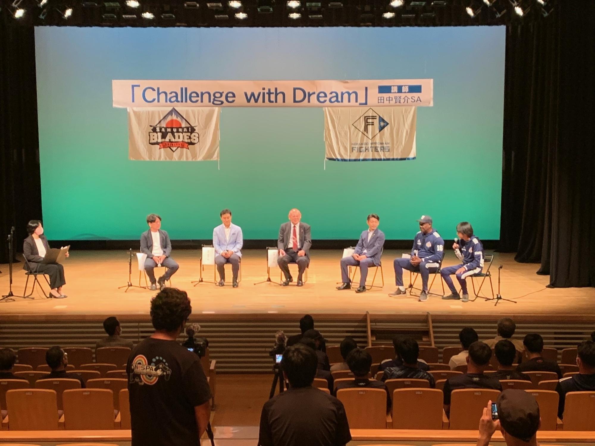 「Challenge with Dream」と書かれた横断幕が掲げられたステージ上に6名の関係者と司会者が座り、トークライブが行われている様子の写真